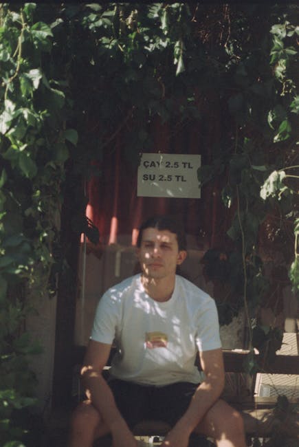 A young man with dark hair and light skin, wearing a white T-shirt and dark shorts, is seated outside in a garden setting. He appears relaxed with his arms resting on his knees. Behind him, there is a backdrop of lush green foliage, including ivy or climbing plants, surrounding a small door or entrance with a fabric curtain. A sign hanging above the door displays prices for Turkish tea and coffee, written in Turkish. The scene is lit naturally with sunlight filtering through the leafy canopy, casting dappled light and shadows across the scene. The garden area shows a combination of soil, plant beds, and paved pathways, indicative of maintained outdoor space, possibly part of a small café or outdoor seating area. Gardeners Harrow might be involved in the upkeep or landscaping of this setting, emphasizing the theme of garden maintenance and outdoor ambiance.