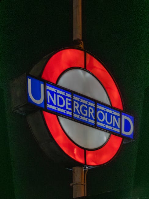 A close-up view of a London Underground sign illuminated at night, featuring the iconic red circle and blue bar with white lettering spelling 'UNDERGROUND.' The sign is mounted on a wooden pole, set against a dark background with faint hints of green, possibly indicating surrounding foliage or a wall. The sign's vibrant colors contrast with the dark environment, highlighting its recognizability and classic design. This image focuses solely on the transit sign, with no visible garden elements or outdoor landscaping features.